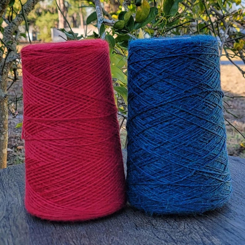 Pode incluir: Dois grandes carret&eacute;is c&oacute;nicos de fio colocados lado a lado. Um carretel &eacute; de uma cor vermelha s&oacute;lida vibrante e o outro carretel &eacute; de uma cor azul s&oacute;lida profunda. Ambos os carret&eacute;is apresentam uma textura de fio fino enrolado.