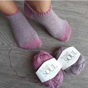 May include: A pair of hand-knitted socks in shades of gray and pink, with matching yarn balls labeled "African Expressions SOUL". The socks have a ribbed cuff and a solid pink toe and heel. The yarn is a soft, textured material.