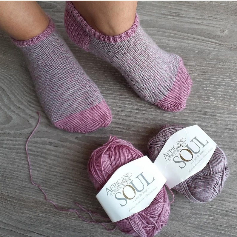 May include: A pair of hand-knitted socks in shades of gray and pink, with matching yarn balls labeled "African Expressions SOUL". The socks have a ribbed cuff and a solid pink toe and heel. The yarn is a soft, textured material.