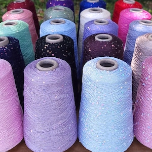 May include: A collection of conical spools of yarn in various colors including lavender, light blue, black, purple, pink, and green. Each spool is wrapped with thread featuring small, reflective sequins distributed throughout the surface.