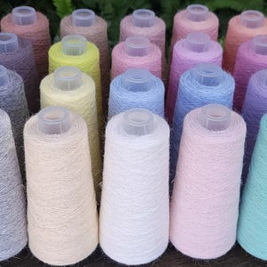 Pode incluir: Uma coleção de vários cones de fio em diversas cores pastel e suaves, incluindo branco, creme, amarelo, rosa, azul e lavanda. Cada cone apresenta um topo de plástico transparente e é envolvido por um material de fio fibroso e texturizado.