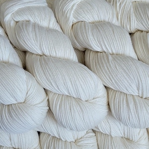 Pode incluir: Close-up de vários novelos de fio branco sujo. O fio é torcido com força, mostrando os fios individuais. A textura do fio é visível, sugerindo uma fibra natural e macia. A imagem é bem iluminada, destacando a cor e a textura do fio.