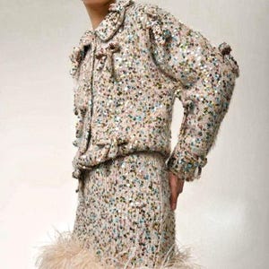 May include: A long-sleeved jacket and matching skirt set crafted from a beige knit fabric densely covered in iridescent, multi-colored sequins. The jacket features a collar, front button closures, and a hemline trimmed with light beige feathers.