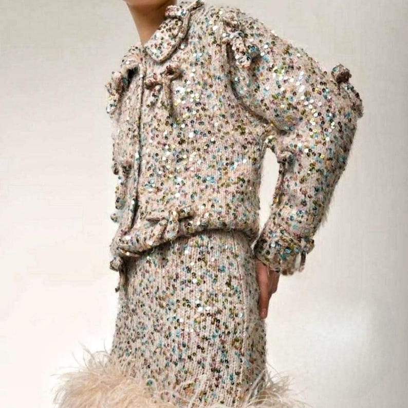 May include: A long-sleeved jacket and matching skirt set crafted from a beige knit fabric densely covered in iridescent, multi-colored sequins. The jacket features a collar, front button closures, and a hemline trimmed with light beige feathers.
