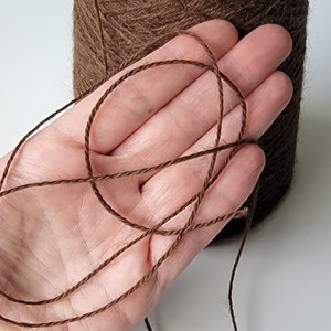 Pode incluir: Um carretel grande de fio castanho escuro com uma apar&ecirc;ncia texturizada e fibrosa. Um &uacute;nico fio do mesmo castanho escuro &eacute; puxado do carretel, exibindo a sua constru&ccedil;&atilde;o torcida de v&aacute;rias camadas e uma cor castanha profunda consistente.