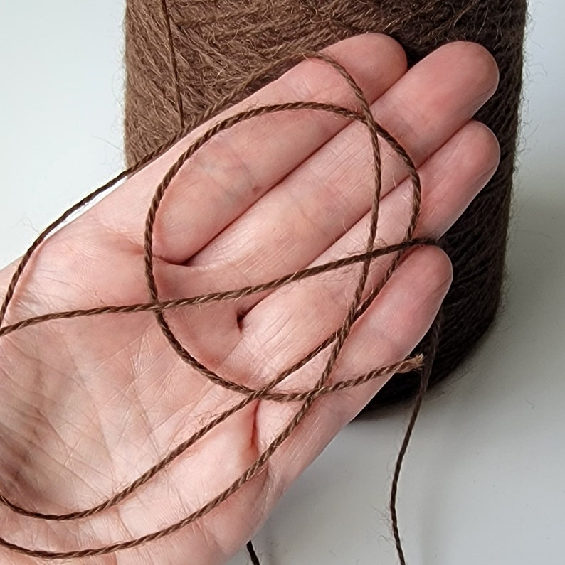 Pode incluir: Um carretel grande de fio castanho escuro com uma apar&ecirc;ncia texturizada e fibrosa. Um &uacute;nico fio do mesmo castanho escuro &eacute; puxado do carretel, exibindo a sua constru&ccedil;&atilde;o torcida de v&aacute;rias camadas e uma cor castanha profunda consistente.
