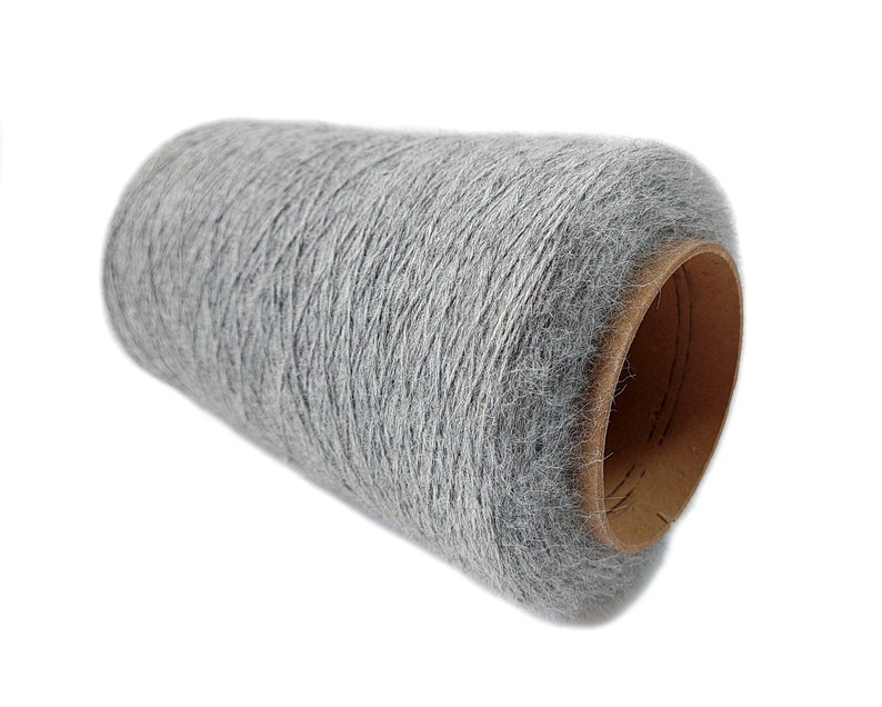 Pode incluir: Um grande cone cil&iacute;ndrico de fio cinza-claro com uma superf&iacute;cie texturizada e felpuda. O fio &eacute; enrolado firmemente em torno de um n&uacute;cleo de papel&atilde;o marrom resistente, exibindo uma apar&ecirc;ncia macia e consistente em toda a bobina.