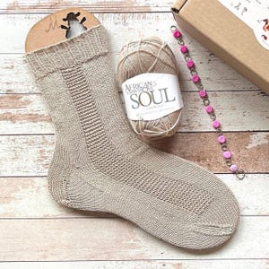 May include: A beige knitted sock with a textured pattern, a ball of yarn labeled "African Expressions Soul", and a pink beaded bracelet. The sock is displayed with a cardboard tag. The items are arranged on a wooden surface.