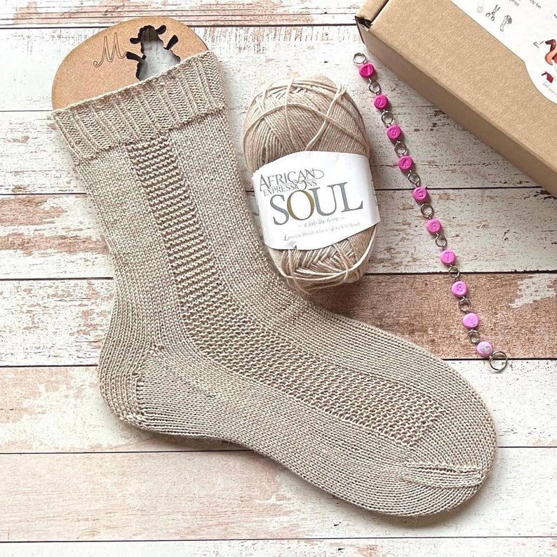 May include: A beige knitted sock with a textured pattern, a ball of yarn labeled "African Expressions Soul", and a pink beaded bracelet. The sock is displayed with a cardboard tag. The items are arranged on a wooden surface.