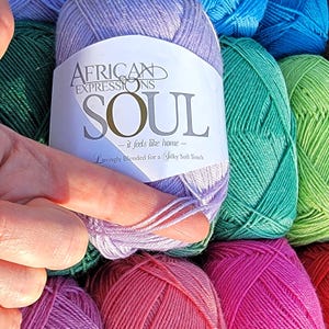 May include: A close-up of a skein of lavender yarn from the "African Expressions Soul" collection. The label reads "Soul - it feels like home - Lovingly blended for a Silky Soft Touch." Other skeins of yarn in various colors are in the background.