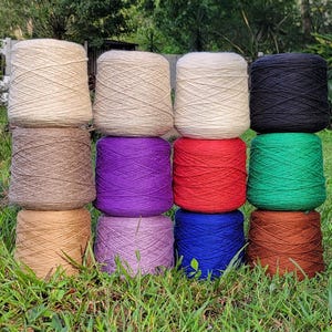 May include: A collection of colorful yarn skeins in various shades, including beige, brown, purple, red, green, blue, and black. The yarn is arranged on a bed of green grass, showcasing the vibrant colors and textures of the fiber.