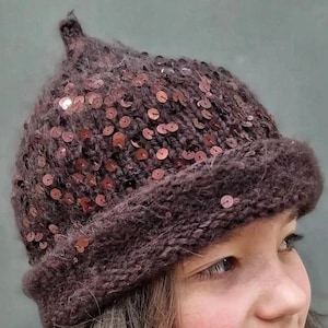 May include: A dark brown knit beanie with a pointed top and a folded brim. The hat is embellished with numerous small, round, dark brown sequins scattered across the surface, providing a subtle reflective texture to the fuzzy, textured yarn.