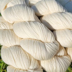 May include: Close-up of off-white yarn skeins. The yarn is thick and twisted, with visible texture. The skeins are arranged in a close, overlapping pattern, suggesting a soft, natural fiber. The background is a blurred green.