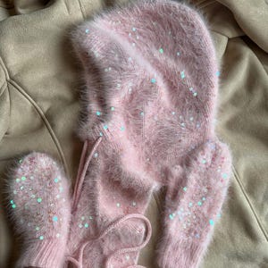 May include: A light pink fuzzy balaclava hood and matching mittens set. The knit fabric features a soft, brushed texture and is embellished with scattered iridescent sequins that reflect light, creating a shimmering effect across the surface.
