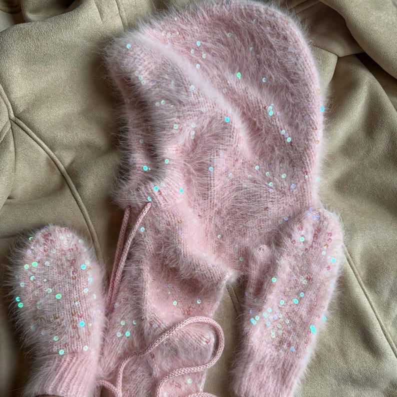 May include: A light pink fuzzy balaclava hood and matching mittens set. The knit fabric features a soft, brushed texture and is embellished with scattered iridescent sequins that reflect light, creating a shimmering effect across the surface.