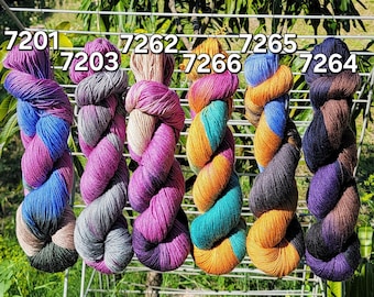 May include: Six skeins of colourful yarn hang from a white rack. The yarn is twisted and features a variety of colours, including purple, blue, grey, teal, orange, and brown. Each skein has a number label: 7201, 7203, 7262, 7266, 7265, and 7264.