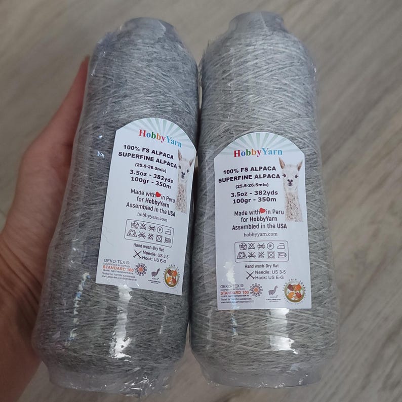 Pode incluir: Dois cones de fio de alpaca superfina cinza-claro embalados em pl&aacute;stico transparente. Cada etiqueta diz HobbyYarn, 100% FS Alpaca, 100g, 350m, 3.5oz, 382yds, com instru&ccedil;&otilde;es de cuidado, agulha US 3-5 e agulha de croch&ecirc; US E-G.