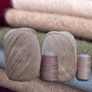 Pode incluir: Dois novelos de lã castanho-claro e dois carretéis de linha menores em tons de taupe e bege, dispostos à frente de várias peças dobradas de tecido de malha macio, felpudo e claro em diversos tons neutros.