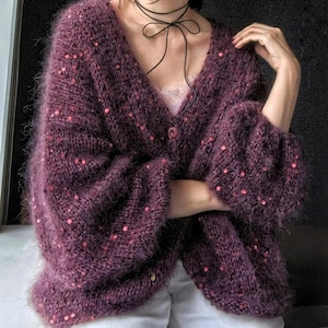 May include: A fuzzy, deep purple V-neck cardigan featuring long, loose sleeves and a single button closure. The knit fabric is embellished with small, scattered red sequins throughout the garment, creating a textured and shimmering appearance.