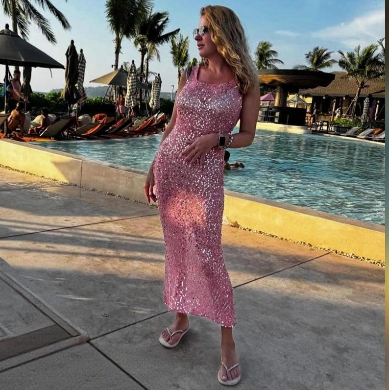 May include: A sleeveless, floor-length dress covered in shimmering pink sequins. The garment features a simple scoop neckline and a straight, form-fitting silhouette that extends to the ankles. The fabric reflects light, creating a bright, metallic appearance throughout the entire surface of the dress.