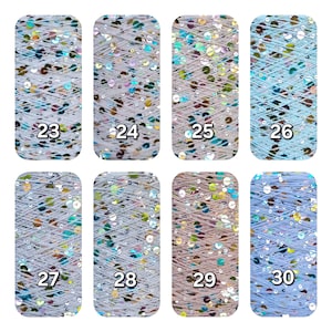 May include: Eight spools of yarn arranged in two rows, labeled 23 through 30. Each spool features light-colored thread interwoven with iridescent, reflective sequins that shimmer in various colors. The spools are rectangular with rounded corners.