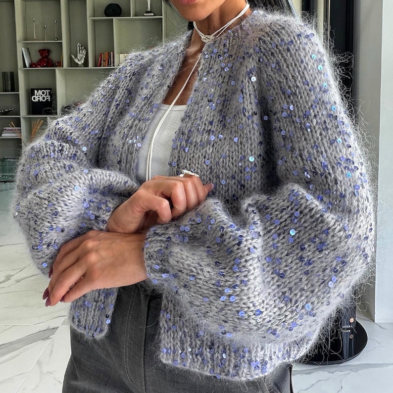 May include: A fuzzy, open-front knit cardigan in a heather gray hue, featuring long, voluminous sleeves and a scattered pattern of small, shimmering light blue sequins throughout the garment, worn over a simple white top and dark gray trousers.