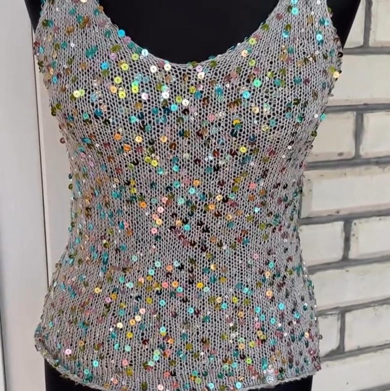 May include: A sleeveless tank top crafted from a light gray knit fabric, featuring a V-neckline and a scattered pattern of small, iridescent, multi-colored sequins in shades of blue, green, pink, and gold throughout the entire garment.