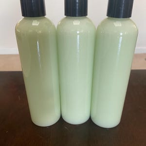 May include: Three clear plastic bottles filled with a pale green liquid, each topped with a black flip-top cap. The bottles are arranged side-by-side on a dark brown wooden surface, suggesting a skincare or cosmetic product.