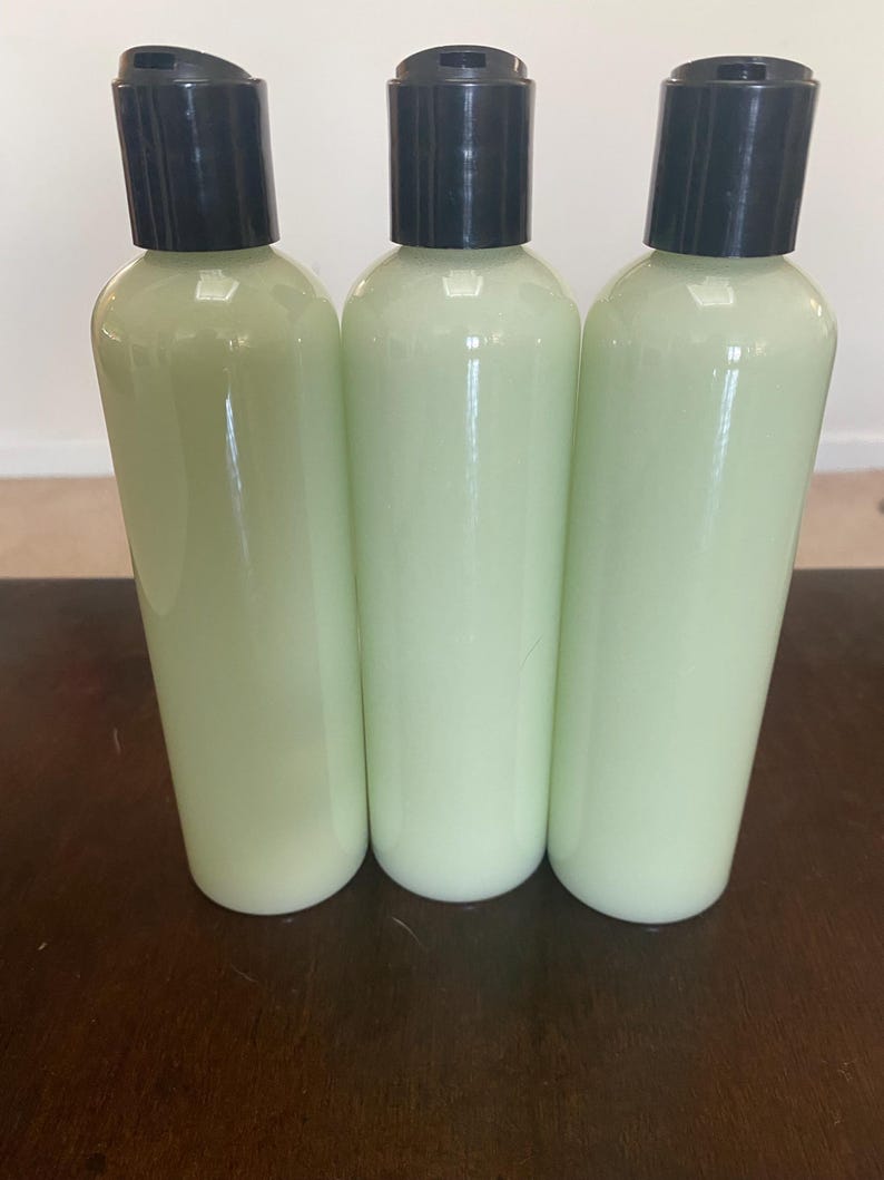 May include: Three clear plastic bottles filled with a pale green liquid, each topped with a black flip-top cap. The bottles are arranged side-by-side on a dark brown wooden surface, suggesting a skincare or cosmetic product.