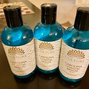 Puede incluir: Tres botellas de pl&aacute;stico transparente con l&iacute;quido azul y una etiqueta blanca que dice "CULTURE Carolina Coastal Breeze Body Wash". La etiqueta tiene un patr&oacute;n geom&eacute;trico dorado y blanco y un logotipo de flor de loto.