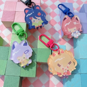 May include: Four acrylic keychains with cartoon character designs. Each keychain features a different character with a unique color scheme, including blue, pink, purple, and yellow. The keychains have metal clasps in black, blue, green, and pink.