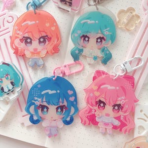 May include: Four acrylic keychains featuring stylized anime-inspired characters with large eyes and colorful hair in shades of orange, teal, blue, and pink. Each charm includes a plastic or metal lobster clasp and displays unique facial expressions.