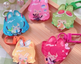 May include: A collection of colorful acrylic keychains featuring anime-style characters. The keychains are in various shapes and colors, including pink, blue, yellow, and green, with attached metal clasps. They are arranged on a wooden tray.
