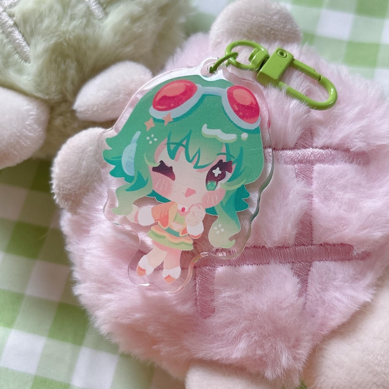 May include: A clear acrylic keychain featuring a cartoon character with green hair, red goggles, and a colorful outfit. The keychain has a green clasp and is set against a pink, fluffy background.