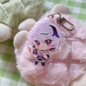 May include: A clear acrylic keychain featuring a cartoon character with purple hair and clothing. The character has large eyes and is holding a pose. The keychain has a silver clasp and is set against a pink, fluffy background.