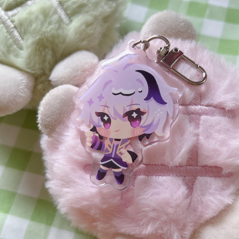 May include: A clear acrylic keychain featuring a cartoon character with purple hair and clothing. The character has large eyes and is holding a pose. The keychain has a silver clasp and is set against a pink, fluffy background.
