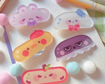 May include: A collection of six colorful, cartoon-style acrylic pins. The pins feature various designs, including a cloud, a popsicle, and a grumpy apple. The pins are in shades of pink, yellow, and purple, with cute facial expressions. Decorative pom-poms and markers are also visible.