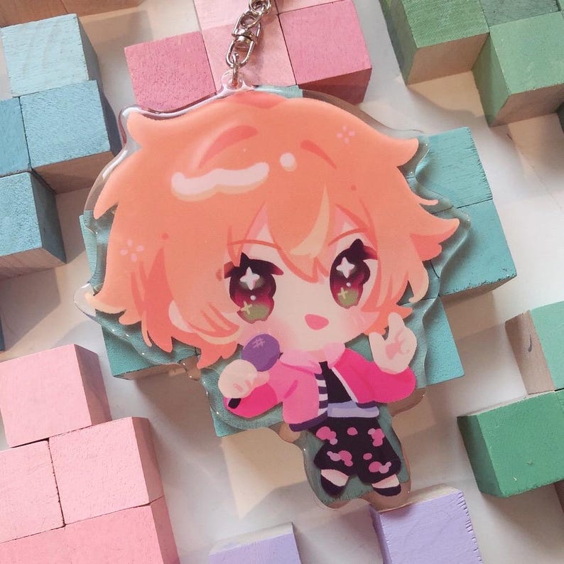 May include: A clear acrylic keyring featuring a cartoon character with orange hair, wearing a pink jacket and black trousers with a microphone. The keyring is attached to a silver chain. The background includes pastel-coloured wooden blocks.