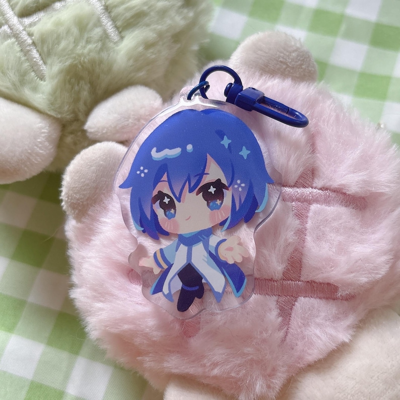 May include: A clear acrylic charm featuring a blue-haired anime character with large eyes and a white and blue outfit. The charm is attached to a blue metal clasp. The background is a soft pink plush material.