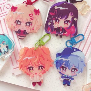 May include: Four acrylic keychains featuring chibi-style anime characters with large expressive eyes. The characters have hair in shades of blonde, dark purple, orange, and blue, each attached to a colorful plastic lobster clasp in red, black, green, and blue.