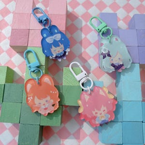 May include: Four acrylic keychains with cartoon character designs. Each keychain features a different color scheme: blue, orange, pink, and light green. The keychains have a lobster clasp for easy attachment to bags or keys. The background is a colorful arrangement of wooden blocks.