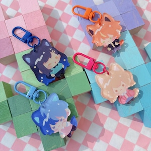May include: Four colorful keychains featuring anime-style characters with fox-like ears. Each keychain has a different color scheme, including blue, orange, and pink, and is attached to a matching carabiner clip. The background includes wooden blocks and a pink and white checkered pattern.