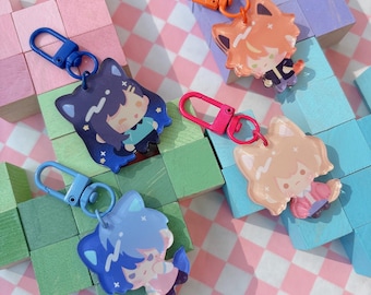 May include: Four colorful keychains featuring anime-style characters with fox-like ears. Each keychain has a different color scheme, including blue, orange, and pink, and is attached to a matching carabiner clip. The background includes wooden blocks and a pink and white checkered pattern.