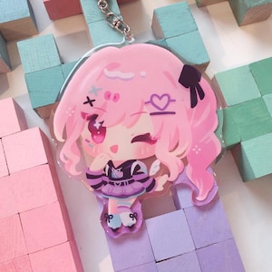 May include: A pink anime-style charm with a character sporting pink hair, a black bow, and a purple and black outfit. The charm has a silver clasp and is set against a backdrop of pastel-coloured wooden blocks.