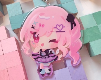 May include: A pink anime-style charm featuring a character with pink hair, a black bow, and a purple and black outfit. The charm has a silver clasp and is set against a background of pastel-colored wooden blocks.