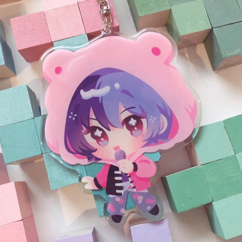 May include: A clear acrylic keyring featuring a cartoon character in a pink bear hoodie. The character has purple hair and is holding a microphone. The keyring has a silver metal ring and clasp.