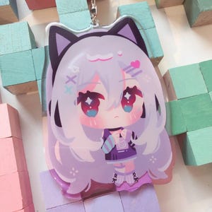 May include: A clear acrylic keychain depicting a cartoon anime character with light purple hair and cat ears. The character has large, sparkling red eyes and wears a purple dress. The keychain has a silver metal clasp.