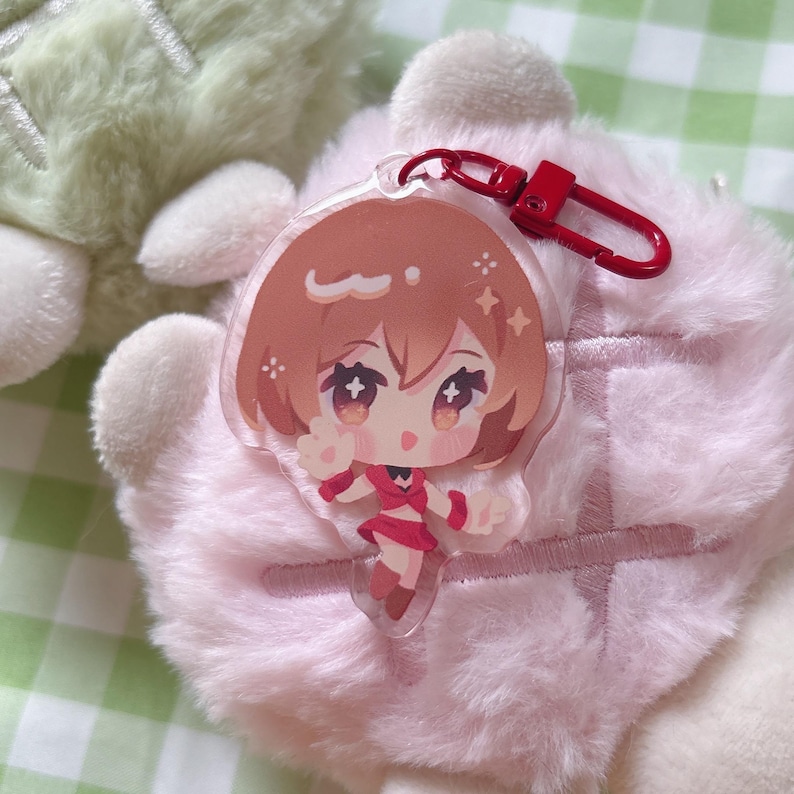 May include: A clear acrylic keychain featuring a cartoon character with brown hair and a red outfit. The keychain has a red clasp and is set against a soft, pink background.