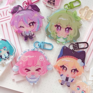 May include: A collection of acrylic keychains featuring various anime-style characters with colorful hair and outfits. The charms include pink, purple, green, and blonde figures with expressive faces, attached to plastic clasps in matching colors.