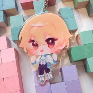 May include: A clear acrylic keychain depicting a chibi anime character with peach-coloured hair and large red eyes. The character is dressed in a white and blue outfit with purple trousers. The keychain has a silver metal ring.